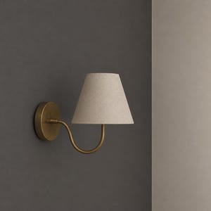 K&ouml;nnte beinhalten: Eine Wandleuchte mit einem kreisf&ouml;rmigen, messingfarbenen Sockel und einem geschwungenen Arm, der einen konischen, hellbeigen Stofflampenschirm tr&auml;gt. Die Lampe ist an einer dunkelgrauen Wand montiert und bietet eine fokussierte Lichtquelle.