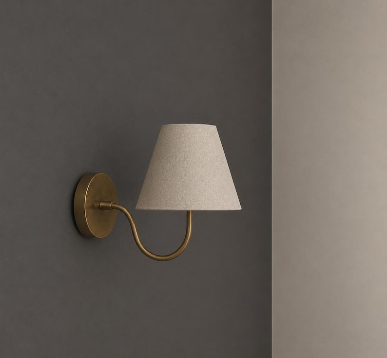 K&ouml;nnte beinhalten: Eine Wandleuchte mit einem kreisf&ouml;rmigen, messingfarbenen Sockel und einem geschwungenen Arm, der einen konischen, hellbeigen Stofflampenschirm tr&auml;gt. Die Lampe ist an einer dunkelgrauen Wand montiert und bietet eine fokussierte Lichtquelle.