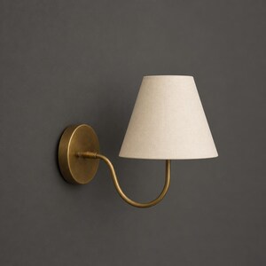 K&ouml;nnte beinhalten: Eine Wandleuchte mit einem kreisf&ouml;rmigen, messingfarbenen Sockel und einem geschwungenen Arm, der einen konischen, cremewei&szlig;en Stofflampenschirm tr&auml;gt. Die Lampe ist so konzipiert, dass sie von der Wand absteht und Licht spendet.