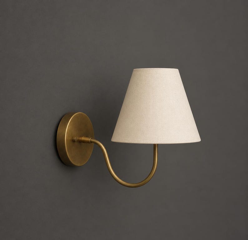 K&ouml;nnte beinhalten: Eine Wandleuchte mit einem kreisf&ouml;rmigen, messingfarbenen Sockel und einem geschwungenen Arm, der einen konischen, cremewei&szlig;en Stofflampenschirm tr&auml;gt. Die Lampe ist so konzipiert, dass sie von der Wand absteht und Licht spendet.
