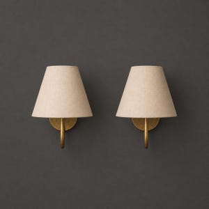 K&ouml;nnte beinhalten: Ein Paar Wandleuchten mit konischen, hellbeigen Stoffschirmen. Jede Lampe wird von einem geschwungenen, messingfarbenen Metallarm getragen, der an einer runden Basisplatte befestigt ist und ein klares, funktionales Lichtdesign bietet.