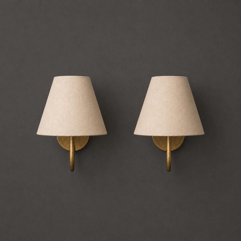 K&ouml;nnte beinhalten: Ein Paar Wandleuchten mit konischen, hellbeigen Stoffschirmen. Jede Lampe wird von einem geschwungenen, messingfarbenen Metallarm getragen, der an einer runden Basisplatte befestigt ist und ein klares, funktionales Lichtdesign bietet.