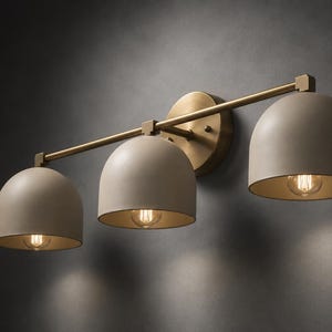 May include: A three-light wall fitting featuring a horizontal brass bar and a circular brass wall mount. Three dome-shaped, matte beige shades are suspended from the bar, each housing a single exposed light bulb that emits a warm glow.