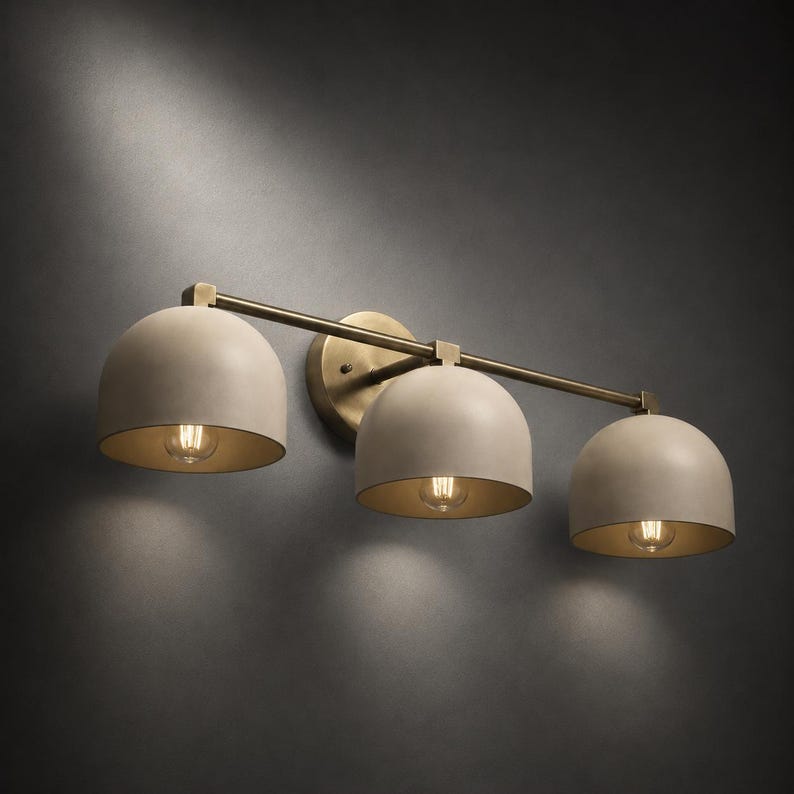 May include: A wall-mounted light fitting featuring three dome-shaped shades in a matte beige finish. The shades are attached to a horizontal brass-toned metal bar and a circular wall plate, each housing a single glowing light bulb inside.