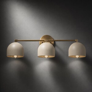 May include: A three-light wall sconce featuring a horizontal brass bar and a circular mounting plate. Three matte beige dome-shaped shades are evenly spaced along the bar, each housing a single light bulb that casts a warm glow downwards.