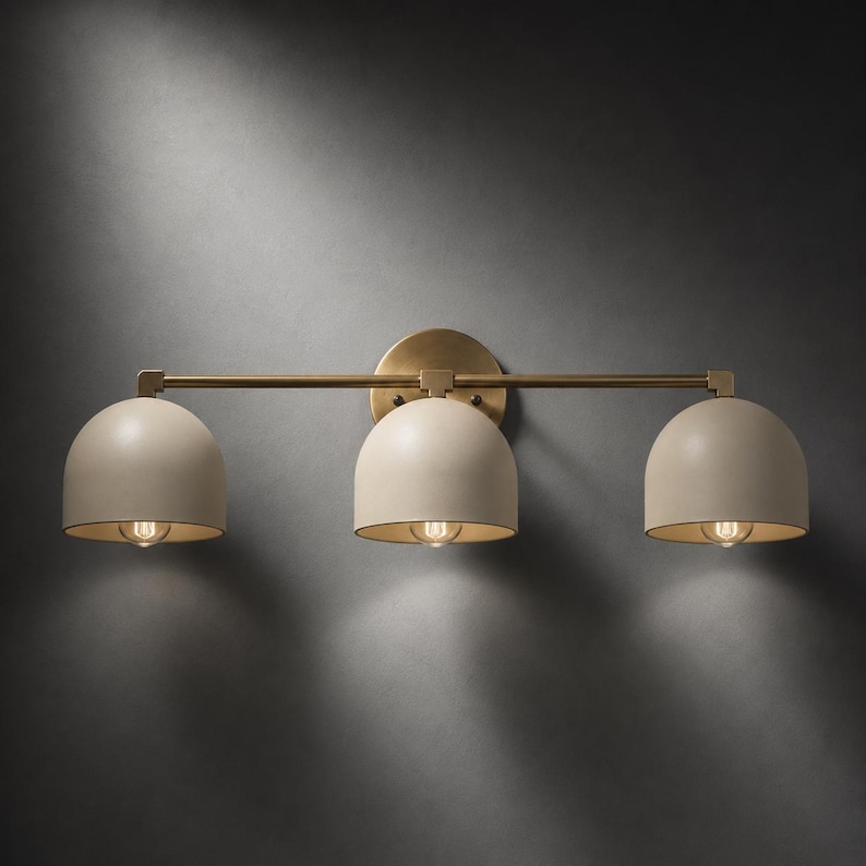 May include: A three-light wall sconce featuring a horizontal brass bar and a circular mounting plate. Three matte beige dome-shaped shades are evenly spaced along the bar, each housing a single light bulb that casts a warm glow downwards.