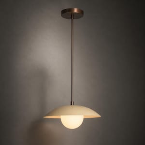 May include: A pendant light fitting featuring a circular, matte cream-coloured metal shade and a central frosted glass globe. The fitting is suspended by a thin, dark bronze-toned metal rod connected to a matching circular ceiling canopy mount.