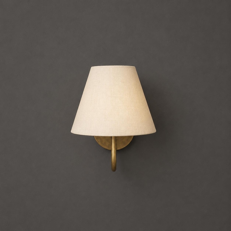 K&ouml;nnte beinhalten: Eine Wandleuchte mit einem konischen, cremewei&szlig;en Stoffschirm, der an einem geschwungenen, messingfarbenen Metallarm befestigt ist. Die Leuchte sorgt f&uuml;r ein sanftes, diffuses Licht und besticht durch ihr schlichtes, funktionales Design.