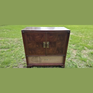 May include: A rectangular wooden cabinet featuring two doors with a dark brown checkerboard wood grain pattern and two vertical brass handles. Below the doors is a horizontal speaker grille covered in a light tan woven fabric with a brass trim.