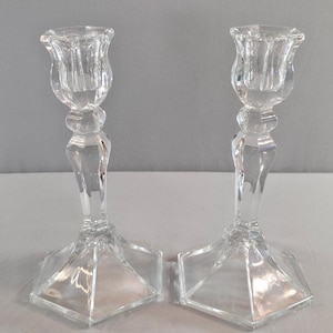 Puede incluir: Un par de candelabros de vidrio transparente con una base hexagonal facetada, un tallo delgado con un nodo decorativo central y una parte superior en forma de tulipán diseñada para sostener una vela cónica. El vidrio es liso y transparente.