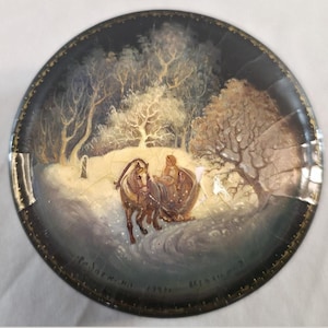 Op de afbeelding: Een ronde zwarte decoratieve schaal met een handgeschilderd wintertafereel van een door een paard getrokken arrenslee in een besneeuwd bos. Het kunstwerk toont een persoon in een slee, kale bomen en een gebarsten lakafwerking. Gouden rand.