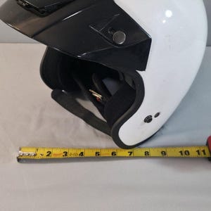 Pode incluir: Um capacete de moto aberto branco com viseira preta e forro interior preto. Uma fita métrica amarela está posicionada à frente do capacete, mostrando uma largura de aproximadamente 28 cm na base do capacete.