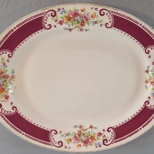Puede incluir: Una bandeja de servicio ovalada de cerámica con un centro de color crema y un borde ancho de color burdeos intenso. El borde está decorado con volutas intrincadas y cuatro grupos de arreglos florales coloridos en rosa, naranja y azul.