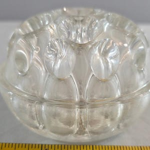 May include: A clear glass flower frog with a dome shape and multiple circular openings for stems. The glass features a textured, molded pattern of rounded indentations across the surface. A yellow measuring tape is visible at the base.