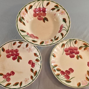 May include: A set of three off-white ceramic plates and a bowl featuring a hand-painted pattern of red cherries, green leaves, and small pink blossoms. Each piece has a scalloped edge and a thin green painted rim, showcasing a consistent floral design.