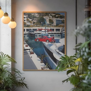 May include: Framed art print featuring a red sports car parked on a yacht in a harbor. The artwork depicts a sunny day with boats and buildings in the background, framed in a gold-colored border.