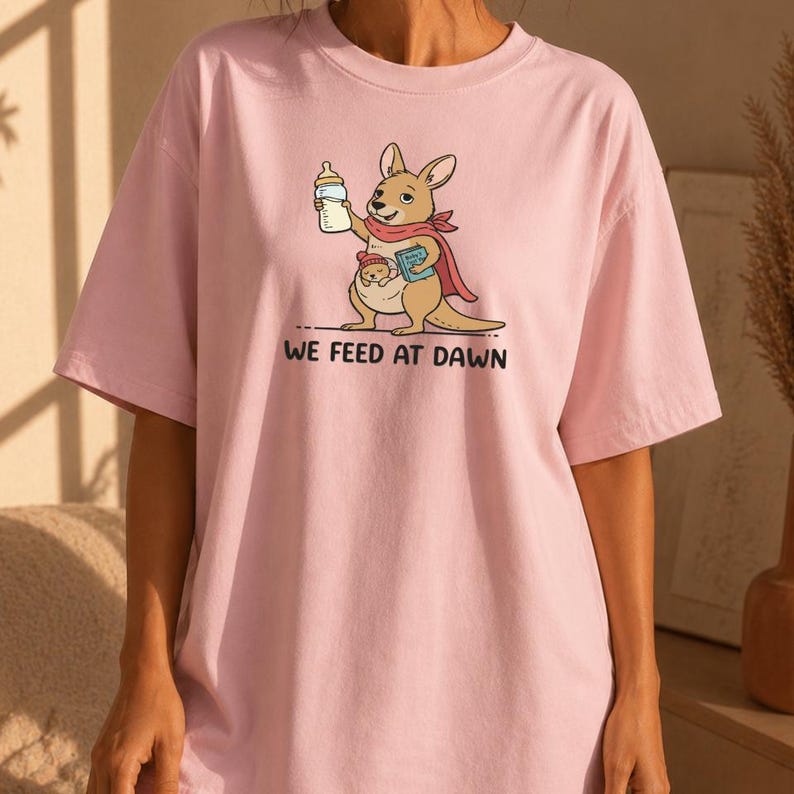 Pu&ograve; includere: Una t-shirt rosa chiaro con la grafica di un canguro marrone che indossa una sciarpa rossa, tiene in mano un biberon e un libro blu, con un piccolo nel marsupio. Il testo "WE FEED AT DAWN" &egrave; stampato in lettere maiuscole nere sotto l'immagine.