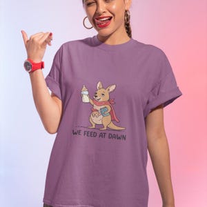 Pu&ograve; includere: Una t-shirt viola a maniche corte con la grafica di un canguro che indossa un mantello rosso, tiene in mano un biberon e un libro, con un piccolo nel marsupio. Il testo "WE FEED AT DAWN" &egrave; stampato in nero sotto l'illustrazione.