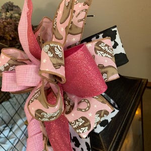 May include: A decorative bow featuring pink and glittery ribbons. The bow incorporates patterned ribbons with cowboy boots, horseshoes, and stars. Accented with a black and white cow print ribbon, this bow is perfect for a country-themed decoration.