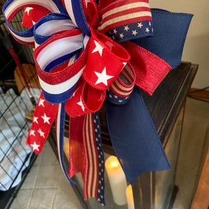 May include: A decorative bow with red, white, and blue ribbons, featuring stars and stripes patterns. The bow is placed on top of a dark lantern with lit candles inside, creating a patriotic theme.