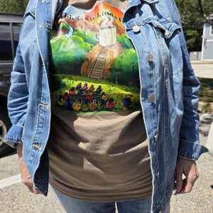 May include: A tan t-shirt featuring a graphic print of a steaming white coffee mug atop a hill with a staircase, surrounded by a group of people, with the text "This is my kinda place" visible at the top, worn under a blue denim jacket.