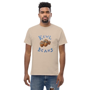 Puede incluir: Una camiseta beige de manga corta con un gráfico de tres granos de café marrones con gafas de sol. El texto "KEWL BEANS" está impreso en letras azules encima y debajo de la ilustración de los granos de café en el centro del pecho.