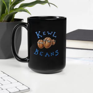 May include: A black ceramic mug featuring a graphic of three brown coffee beans wearing sunglasses. The text "KEWL BEANS" is printed in blue letters above and below the coffee beans. The mug has a curved handle on the left side and a smooth finish.