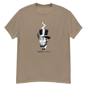 Puede incluir: Una camiseta de manga corta color canela con un gráfico en blanco y negro de una persona con barba y pajarita que lleva un sombrero de copa que parece una taza de café con vapor. El texto ABREWham Lincoln está impreso bajo el gráfico.