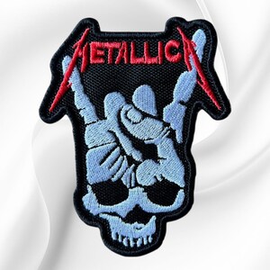 May include: An embroidered patch featuring a light blue skull base with a hand making a rock and roll gesture above it. The word METALLICA is stitched in red thread across the top, with the first and last letters extending into red lightning bolts.