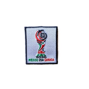 May include: A rectangular embroidered patch with a light gray background and a dark blue border. The center features a stylized trophy graphic in red, green, and black with the text 2026. Below the trophy, the text reads MEXICO USA CANADA.
