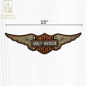 May include: A wide, embroidered Harley-Davidson patch featuring a central shield with the text MOTOR HARLEY-DAVIDSON CYCLES and large, outstretched wings in tan, orange, and black. The patch measures 15 inches in width as indicated by the graphic.