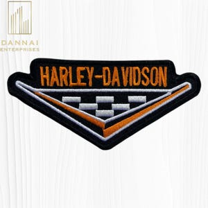 May include: A triangular embroidered patch featuring the text HARLEY-DAVIDSON in orange thread on a black background. Below the text is a white and black checkered pattern bordered by a white and orange V-shaped line on a black fabric base.