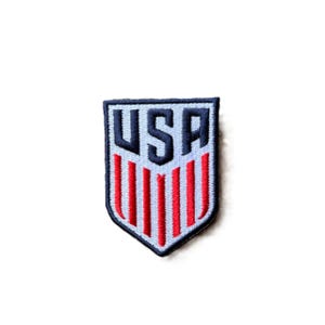 May include: A shield-shaped embroidered patch featuring the letters USA in dark blue at the top, positioned above a series of vertical red stripes on a light blue background, all enclosed within a dark blue border with a pointed bottom edge.