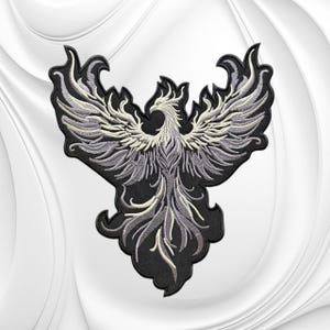 May include: An embroidered patch featuring a stylized phoenix with wings spread wide. The design uses shades of silver, grey, and white thread against a solid black background, creating a high-contrast image of a mythical bird in flight.