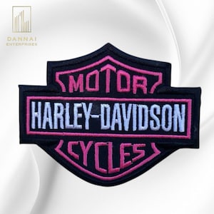 May include: A black embroidered patch in the shape of a shield featuring the text MOTOR in pink, HARLEY-DAVIDSON in white, and CYCLES in pink. The design is outlined with pink stitching and has a black background with a distinct bar and shield shape.