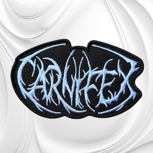 May include: An embroidered patch with a black background and light blue, stylized, jagged lettering that reads CARNIFEX. The patch has a distinct, irregular oval shape with a black stitched border, featuring intricate, branch-like embroidery.