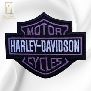 May include: A black embroidered patch in the shape of a shield featuring the text MOTOR HARLEY-DAVIDSON CYCLES. The word HARLEY-DAVIDSON is stitched in white thread, while the words MOTOR and CYCLES are stitched in a light purple thread.