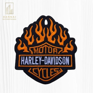 May include: A black embroidered patch featuring orange flame designs at the top. The center contains the text MOTOR in orange, HARLEY-DAVIDSON in white, and CYCLES in orange, all enclosed within a shield-shaped border with orange outlines.