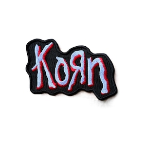 May include: An embroidered patch featuring the word Korn in a stylized, jagged font. The lettering is light blue with a bold red outline, set against a solid black background with a thick, irregular black border following the shape of the text.