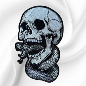 May include: An embroidered patch featuring a detailed human skull in light blue and white thread with a black border. A coiled snake with textured scales is depicted emerging from the open jaw of the skull, rendered in matching monochromatic tones.