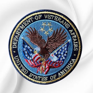 May include: A circular embroidered patch featuring the Department of Veterans Affairs seal. It displays a brown eagle with wings spread, two American flags, and five gold stars on a light blue background, all bordered by dark blue text.