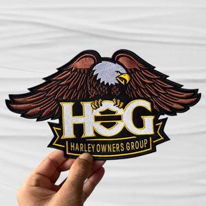 May include: An embroidered patch featuring a bald eagle with brown wings and a white head, positioned above the text HOG in large white letters with a yellow outline. Below, a yellow banner contains the text HARLEY OWNERS GROUP in black.