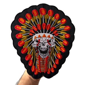 May include: A black embroidered patch featuring a central white skull wearing a vibrant headdress with red and orange circular accents. The skull has red markings around the eyes and a detailed jaw, set against a scalloped black fabric background.