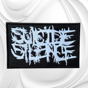 May include: A rectangular embroidered patch with a black background and light blue thread forming the text Suicide Silence in a jagged, stylized font. The patch features a dense, textured stitch pattern throughout the design with a black border.