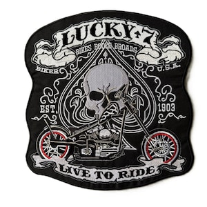 May include: A black embroidered patch featuring a skull centered within a spade shape. The design includes a motorcycle, the text LUCKY 7, BIKES BOOZE BROADS, BIKER, U.S.A., EST. 1903, and LIVE TO RIDE. The motorcycle wheels have red accents.