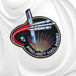 May include: A circular embroidered patch with a black background featuring a white spacecraft, a red curved trail, and a colorful sunburst pattern. The text PROJECT HAIL MARY is stitched in white along the bottom edge, accompanied by two small white stars.