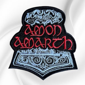 May include: A black embroidered patch featuring the text Amon Amarth in red thread. The design includes a light blue, intricately detailed hammer motif positioned above and below the text, with a black border surrounding the entire shape.