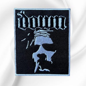 May include: A rectangular embroidered patch with a black background and a white border. The patch features the word "Down" in a stylized gothic font at the top, with a white abstract graphic of a figure wearing a crown of thorns below the text.