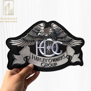 May include: A black embroidered patch featuring a silver eagle with wings spread wide above a motorcycle wheel and the letters HOG. Below the wheel, a silver banner displays the text HARLEY OWNERS GROUP in bold, dark lettering.