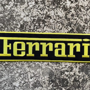 May include: A rectangular black patch with the word "Ferrari" embroidered in yellow. The patch has a yellow border and is set against a textured gray background.